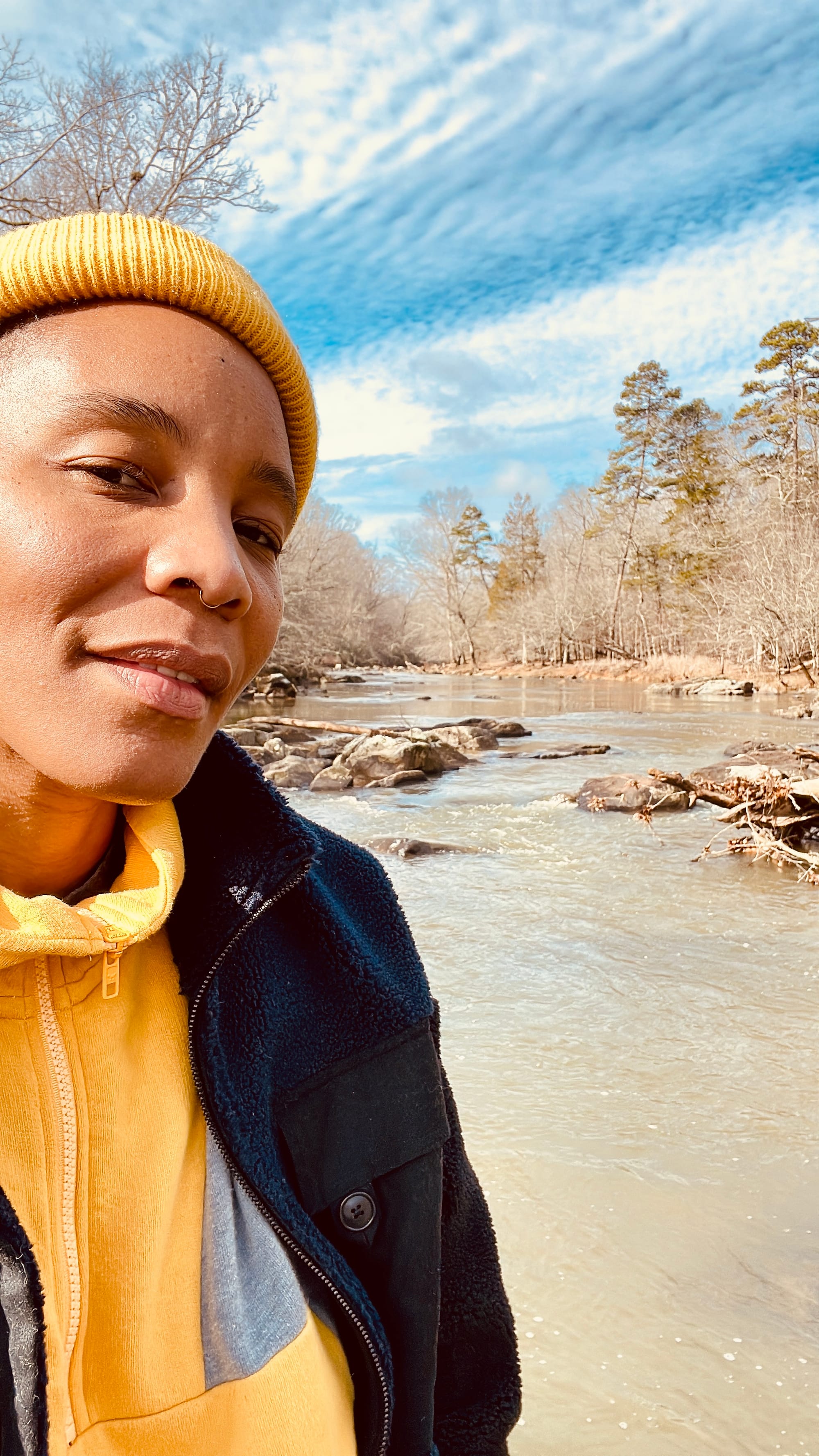 V Woods stands alonside a winter river in a fleece and yellow beanie 